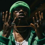 Young man in green jacket and knit cap showcasing jewelry at night with urban backdrop.