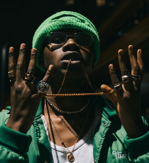 Young man in green jacket and knit cap showcasing jewelry at night with urban backdrop.